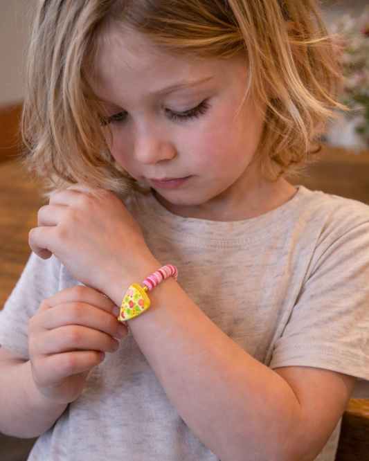 A young child with blonde hair looking down at a colorful heart-shaped bracelet with pink beaded straps, wearing a light gray t-shirt.