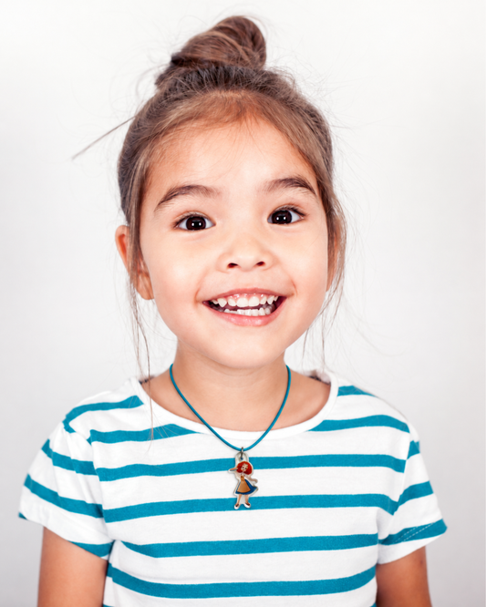 A young girl with a messy bun smiling, wearing a blue and white striped t-shirt and a blue cord necklace with a small character pendant, against a plain white background.