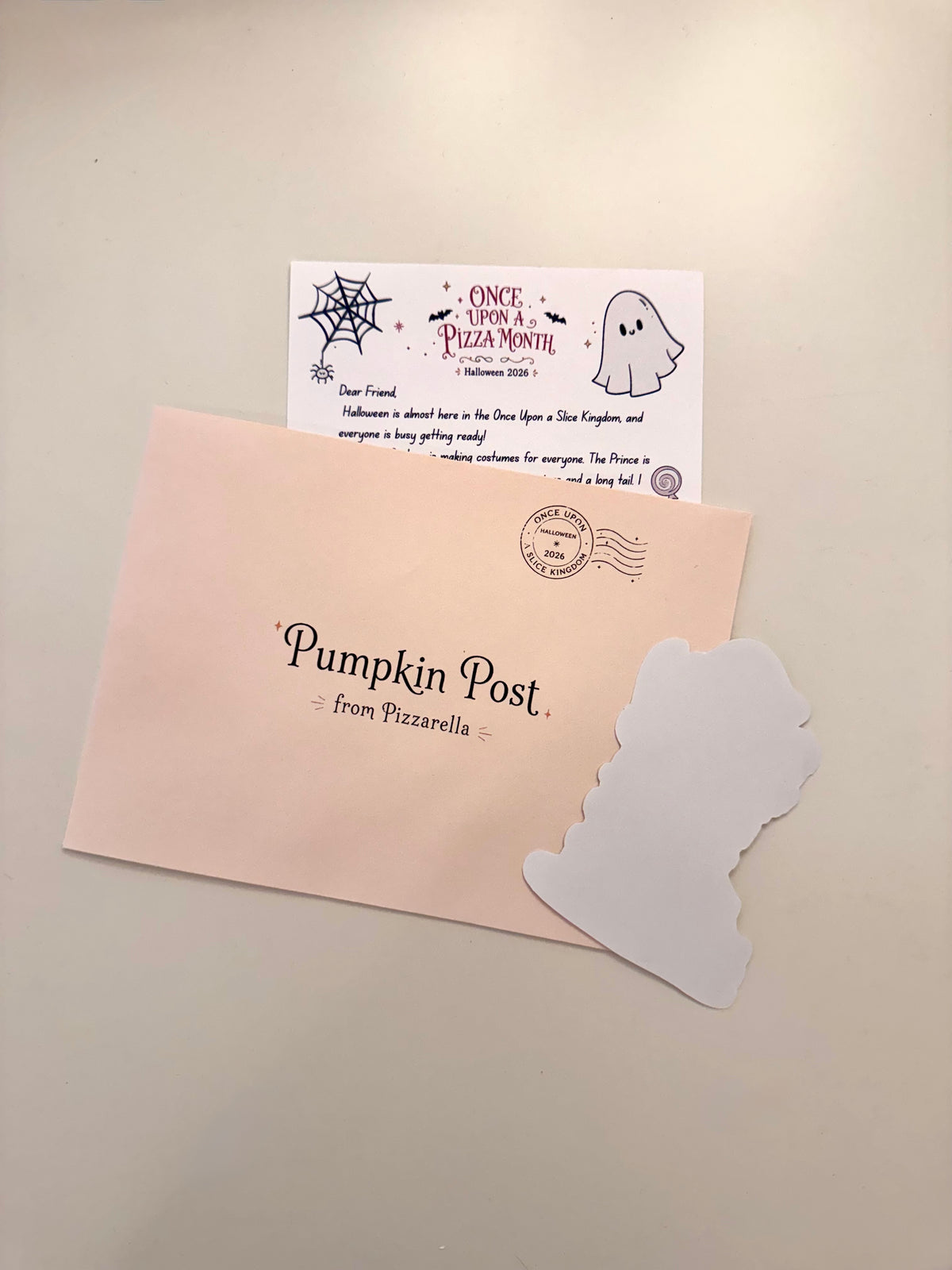 A Halloween-themed postcard set on a light beige surface, featuring a pale pink envelope printed with 'Pumpkin Post from Pizzarella,' a white ghost-shaped card, and a white postcard insert with a ghost illustration and message.