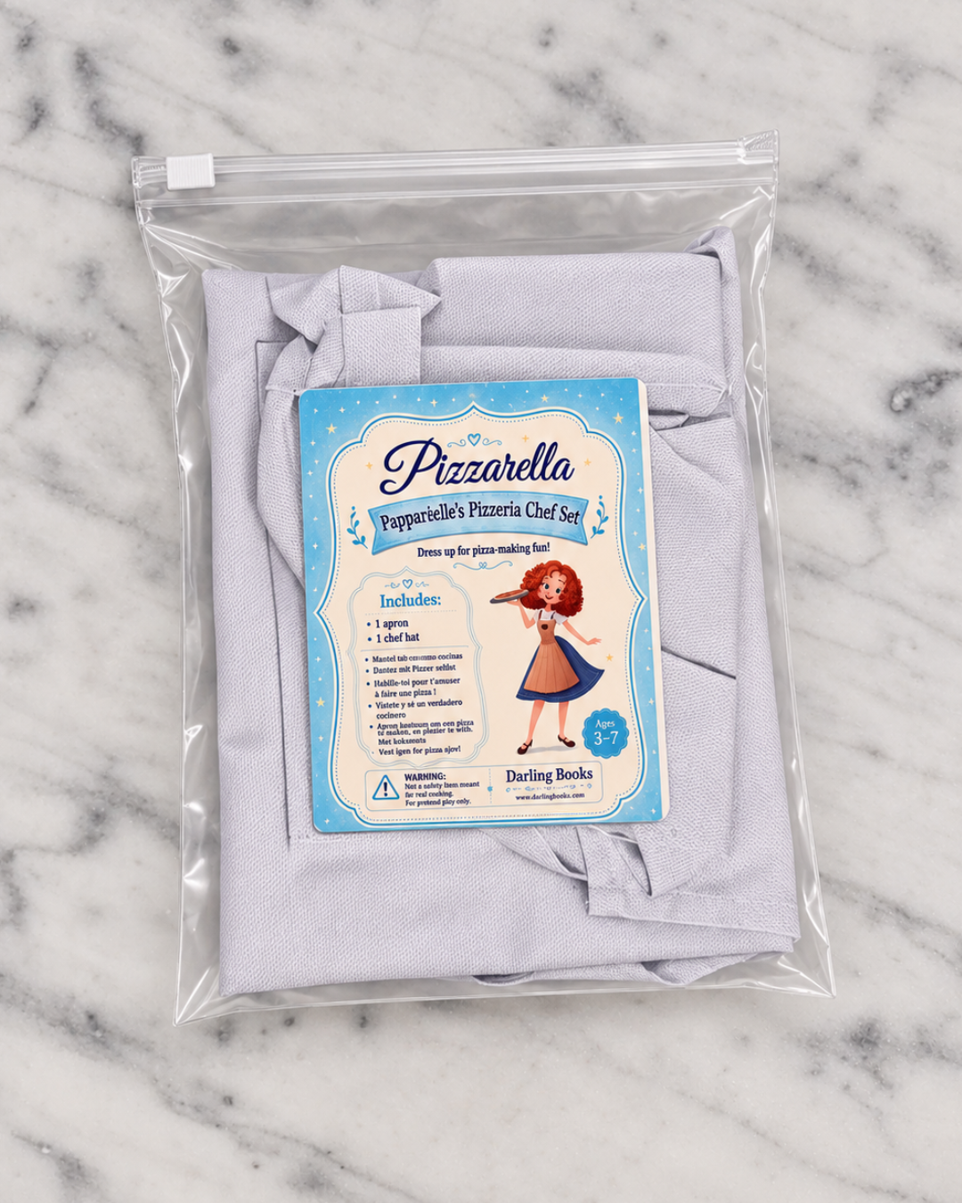 A packaged fabric cooking apron set labeled 'Pizzarella Pepperoni's Pizza Chef Set' in a clear zip bag, placed on a white marble surface. The apron inside is light lavender, and the front label shows a cartoon chef and included items.