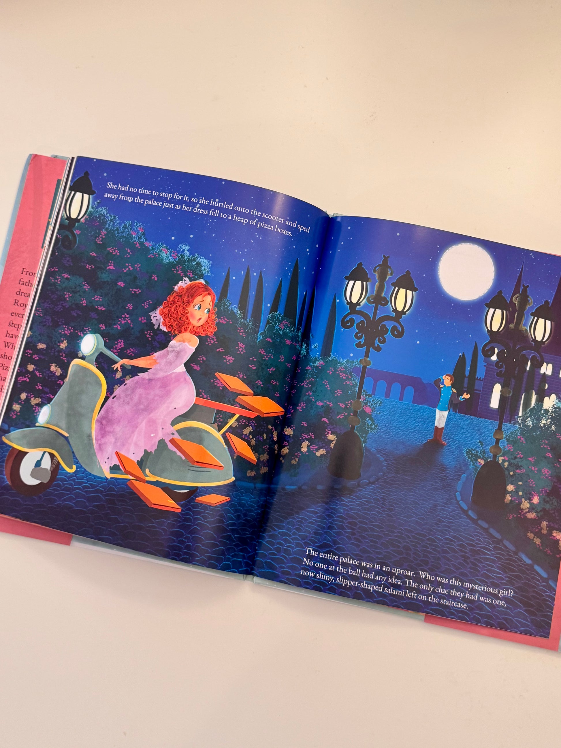 An open children's picture book showing a colorful illustrated night scene with a girl in a pink dress on a scooter and a boy walking under a full moon, photographed on a light beige surface.