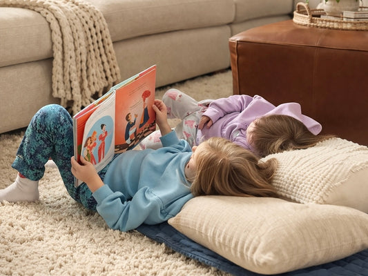 Two children lying on a rug, one reading a book to the other.